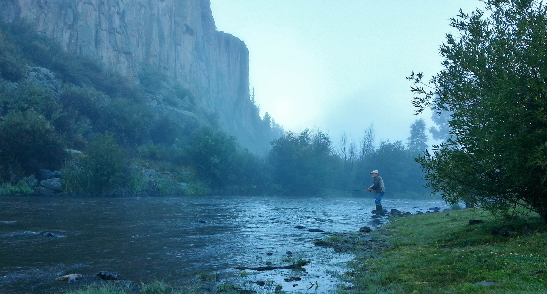 Man fishing in river with green trees and grass, tall cliffs, foggy atmosphere