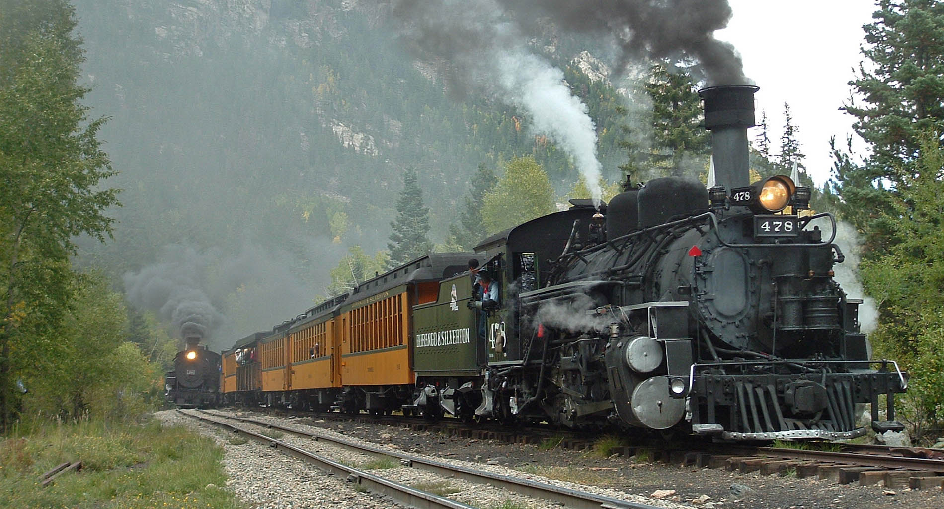 Two old locomotives with black smoke on train tracks with green forest and mountains in background