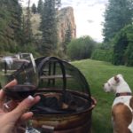 A manicured hand holds a wine glass, sitting near the firepit while looking out at the river with her dog