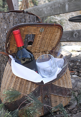 A tan and brown picnic basket with a bottle of wine and two glasses sits on the ground