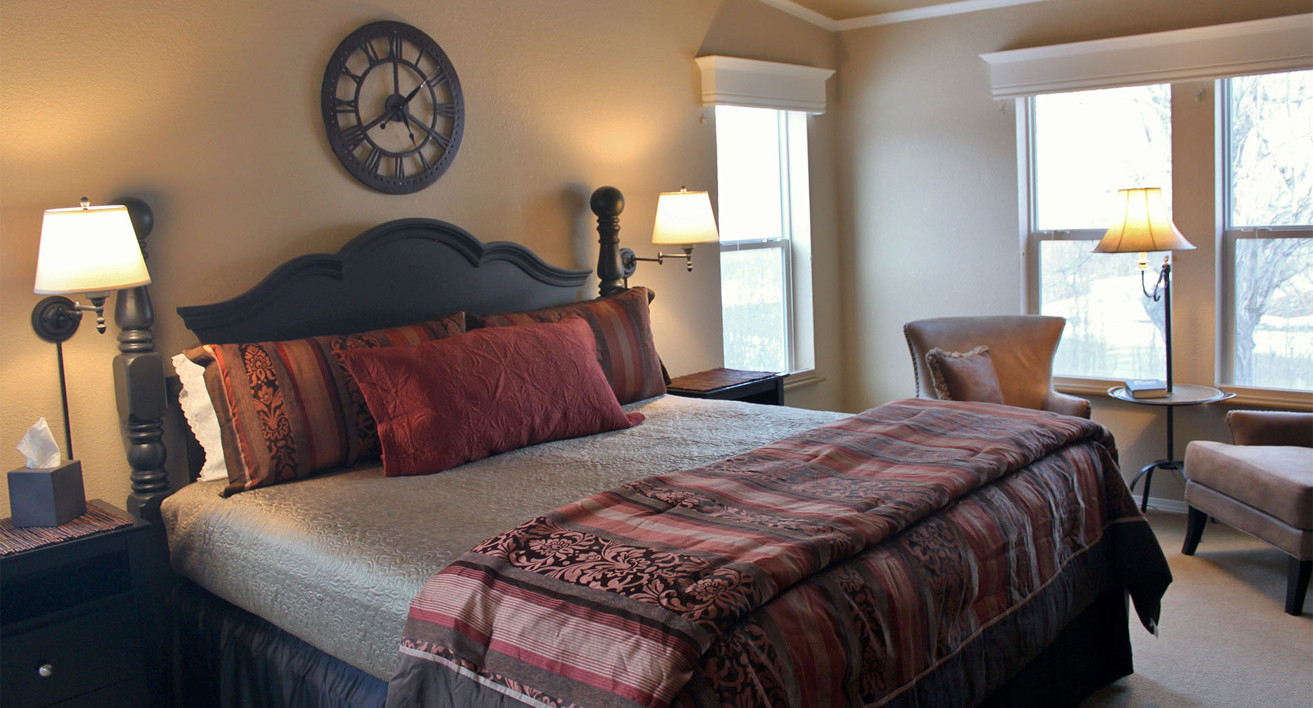 Guestroom with dark wood poster bed and burgandy bedding , with small sitting area near windows