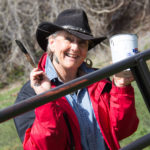 A smiling woman in a red coat and black cowboy hat holds a paintbrush and container of paint