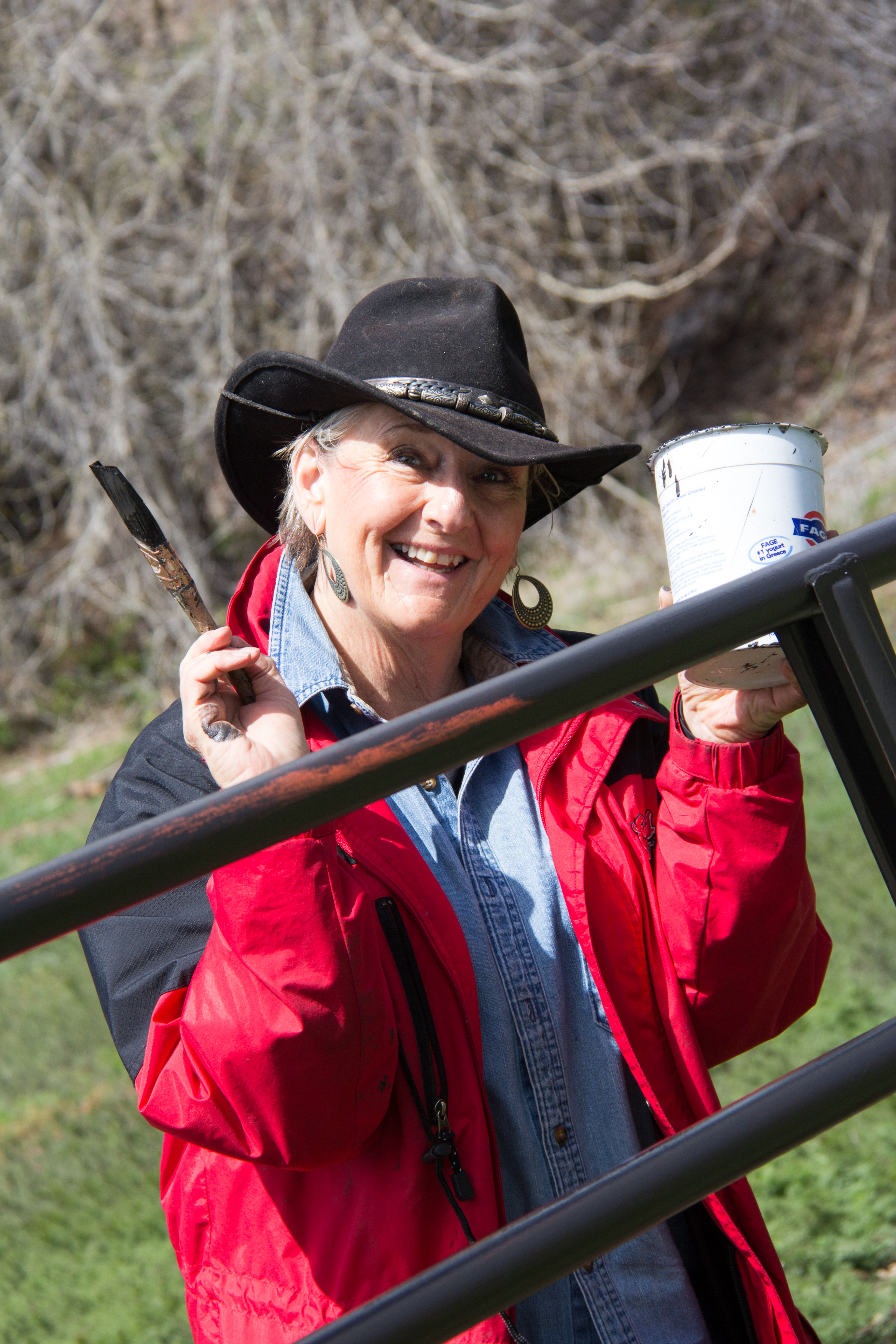 A smiling woman in a red coat and black cowboy hat holds a paintbrush and container of paint