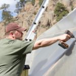 A man in green shirt is standing on a ladder using a drill to build a greenhouse