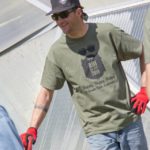 A man in a black hat green shirt and jeans stands with hand on hip smiling