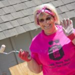 A smiling woman in a pink shirts waves with one hand while holding a paint roller in the other