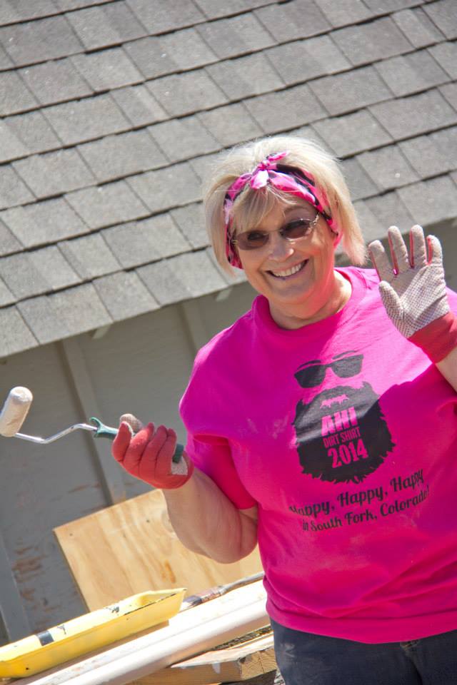 A smiling woman in a pink shirts waves with one hand while holding a paint roller in the other