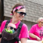 Two smiling ladies, one holding a baby in carrier, are wearing bright pink tshirts and dark sunglasses